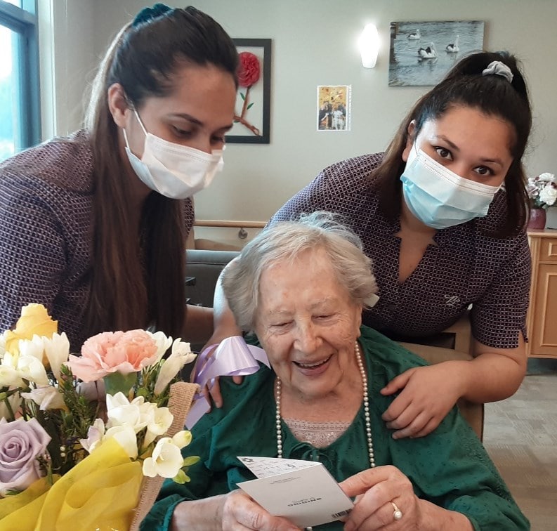 Nada reads her birthday card on her 93rd birthday, flanked by two carers and a huge bouquet of flowers
